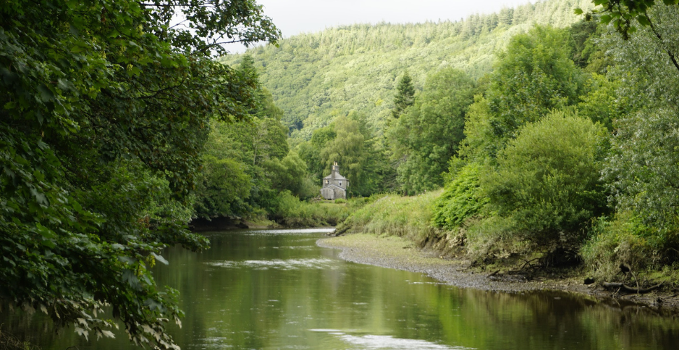 River winding through a lush green valley with dense trees on both banks and a small stone house nestled in the distance.